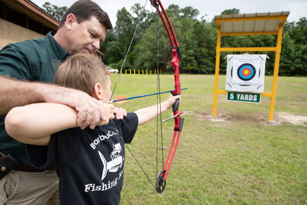 Image of a man helping a child shoot a bow and arrow at a target.