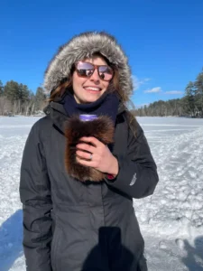 Photo of a woman with her hood up holding a drink that is in beverage koozie made from a beaver pelt.